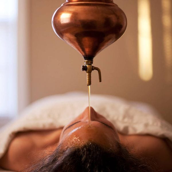 Close-up of an Ayurvedic oil treatment at Sai Ayush Ayurveda Hospital in KPHB, Hyderabad. A copper vessel drips warm oil onto a person's forehead, depicting traditional Shirodhara therapy.