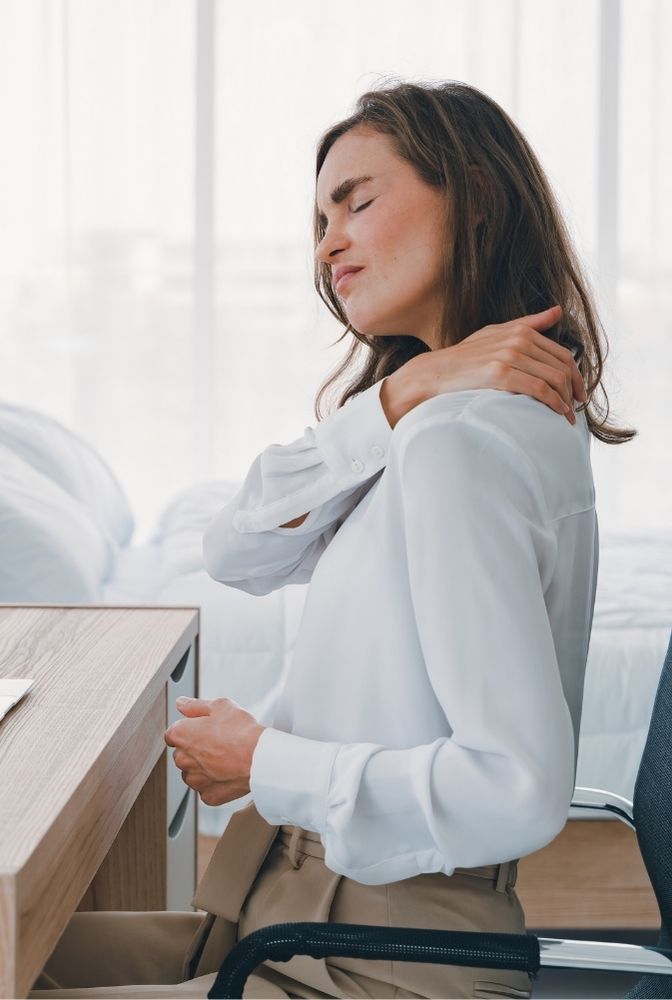 Woman sitting at a desk, wearing a white blouse, experiencing neck pain and massaging her neck. Indicative of seeking Ayurvedic treatment for relief, highlighting herbal therapy and yoga practices in Hyderabad.
